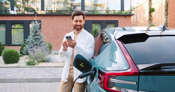 Handsome Caucasian man connecting a car charger to his electric vehicle while using a mobile app on his smartphone to start the charging process