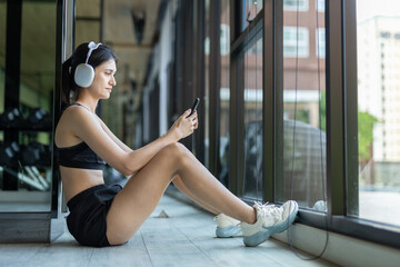 Fit women sitting on gym floor by window, listening to music with headphones and using smartphone. Concept of post workout relaxation, digital lifestyle, fitness tracking and health routine.