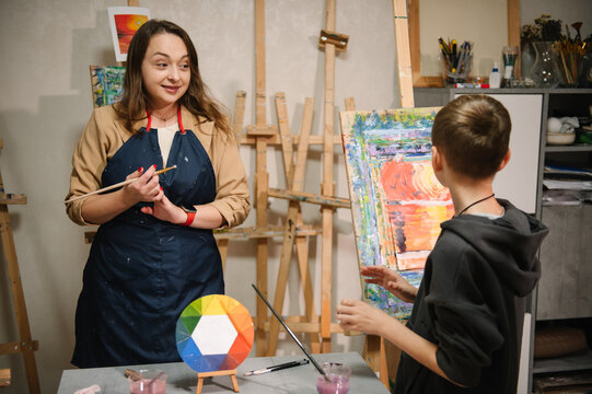 A school boy drawing brush landscapes on easel. Teacher explains to kid how to draw pictures in art class. Child enjoys painting in workshop. Art lesson. Education, creativity, learning concept. - Powered by Adobe