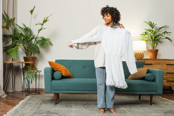 Shopping fashion style. African woman choosing dress at home. Young woman trying to choose dress to wear holding hanger with clothes. Curly dark skinned girl smiling holding hanger with white dress
