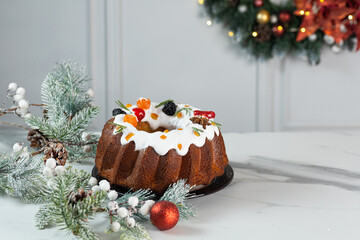 Christmas food still life with iced fruit cake, holly, snow covered winter greenery on light background.