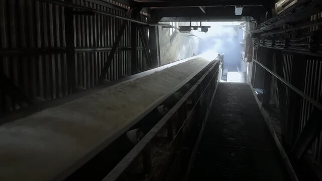 A dynamic, close-up shot captures a heavy industrial conveyor belt transporting a high volume of crushed material, such as cement or limestone, at great speed through a dark factory interior. The stee