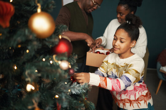Black girl decorating Christmas tree with ornaments while middle aged Black man and Black woman preparing decorations in background, family celebrating holiday together indoors
