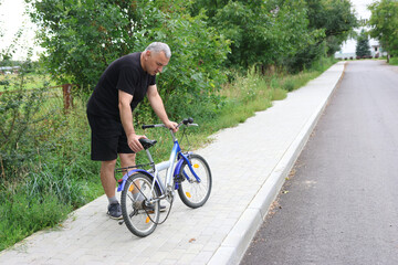 Man in black clothes checking broken chain on a kids bicycle on sidewalk near green fence.
