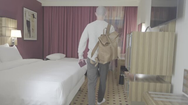 Young man entering hotel room, walking toward window, jet lag body&rsquo;s internal clock, traveler arriving business or vacation stay, feeling relief, comfort after long journey, modern lifestyle solo trip