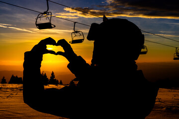 Silhouette of a snowboarder forming a heart shape with hands at sunset on a ski lift