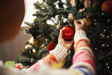 Caucasian child decorating Christmas tree by hanging red ornament, hands visible adjusting bauble among branches, festive lights glowing in background, holiday celebration scene