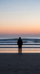 Fototapeta premium Silhouette of a person on a quiet beach, contemplating life while gazing at the tranquil ocean horizon during a beautiful twilight glow