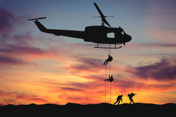 Silhouettes of soldiers descend from a helicopter against the backdrop of sunset. The concept of a military special operation, a special forces mission, and teamwork in extreme conditions. © Andrew L