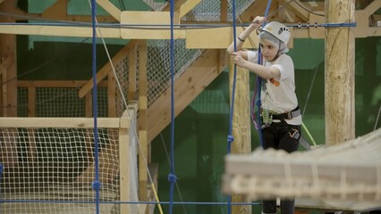 Young girl carefully climbing rope bridge in indoor adventure park. Creative