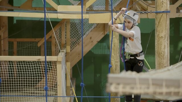 Young girl carefully climbing rope bridge in indoor adventure park. Creative