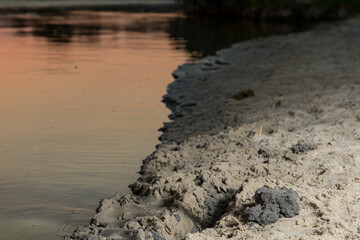 Wet sandy riverbank with clay and small ripples at sunset. Natural detail of shore with soft light reflections.