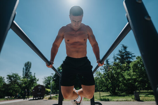 Athletic man practicing strength and balance exercises on parallel bars under clear blue sky, surrounded by vibrant greenery during a sunny day in an outdoor park environment.