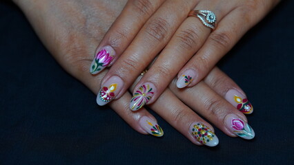 A close-up of a woman's hand featuring festive Diwali-themed nail art. The design includes traditional Indian elements such as diyas, rangoli patterns, glitter, and vibrant colors like gold, red, and 
