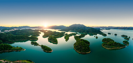 Aerial view over a vast tranquil lake with countless green forest islands under a clear sunrise sky in Qiandao Lake, China.