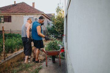 Two men stand by a wheelbarrow filled with branches, trimming garden waste in a suburban yard. They wear gloves and casual clothes, focused on outdoor cleanup near a white-walled house.