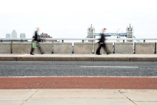 London Rush Hour: Business commuters, blurred by motion, walking along London Bridge with Tower Bridge in the background. The long exposure captures the speed and anonymity of city commuters.