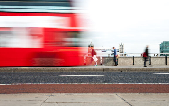 London Rush Hour: A fast-moving red London bus speeds past pedestrians on a pavement near the Thames, with Tower Bridge visible in the background. The motion blur captures the hectic pace of the city.