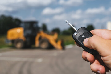 A close-up of a man opening a construction vehicle with a key fob, with a yellow forklift parked in...