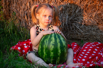 Little girl enjoys summer day with large watermelon while sitting on colorful blanket near hay bales