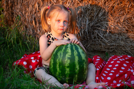 Little girl enjoys summer day with large watermelon while sitting on colorful blanket near hay bales