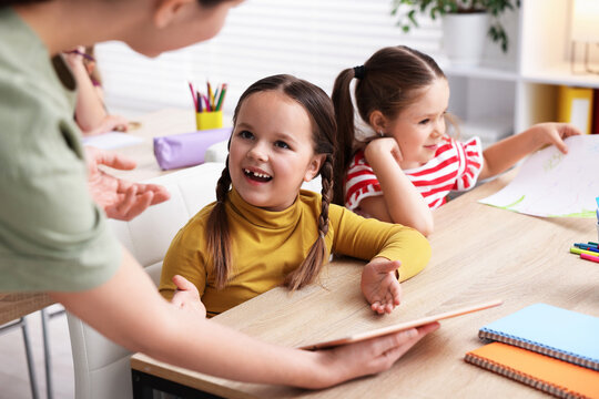 Teacher and smiling little girl with tablet at wooden desk in elementary school, closeup