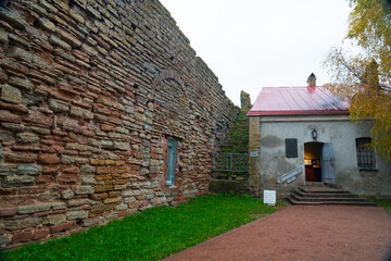 The imposing, rough-hewn stone wall of a fortress next to a small auxiliary building with a red roof, a dirt path, and a tree displaying autumn foliage at Oreshek Fortress