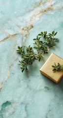 A small gold gift box rests on a pale teal marble surface, adorned with a sprig of fresh green herbs
