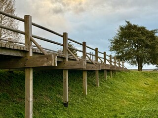 Obraz premium Long wooden footbridge with railings supported by vertical posts over a grassy slope under a cloudy sky.
