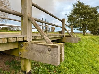 Fototapeta premium Close-up view of a wooden bridge structure with support beams and railings over green grass under a cloudy sky.