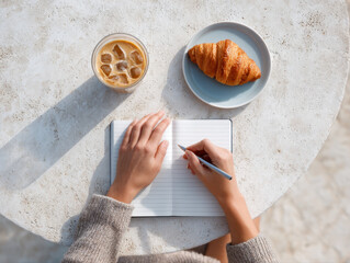 Top down view of person journaling at outdoor table with iced coffee and croissant on plate, enjoying peaceful moment