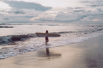Surfer carrying a longboard steps out of gentle waves at dusk. Wet sand mirrors pastel clouds as sea foam curls around her ankles, highlighting gear, balance, and a calm, tech-free coastal routine