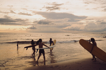 Group of surfers gather at the water’s edge, boards under arm, preparing to paddle at twilight. Golden reflections and layered clouds frame a moment of readiness centered on gear, balance, and intent