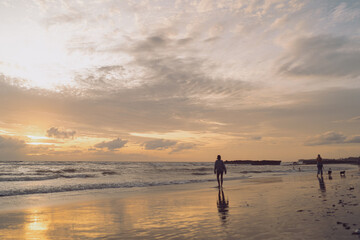 Fototapeta premium People walking on the wet beach at sunset, reflections glowing on the sand, capturing harmony between motion and stillness, presence, and calm connection to nature’s rhythm