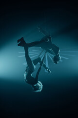 Fototapeta premium Two female aerialists hang inverted on steel chains under blue stage lighting—duo acrobatics in silhouette; vertical frame.