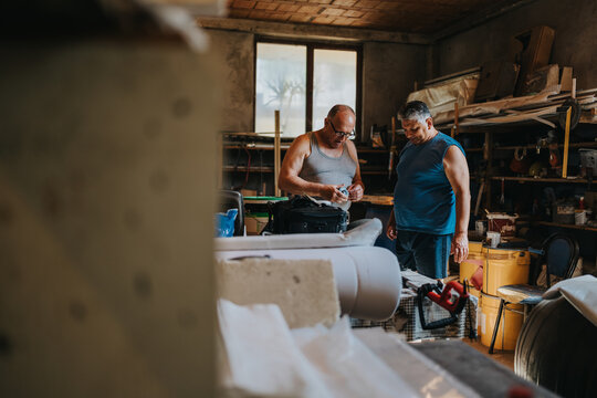 Two men in sleeveless shirts work in a cluttered workshop filled with tools, shelves, and materials. They inspect a piece of equipment at a workbench as natural light fills the space.