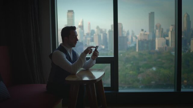 A man dressed in a stylish suit is engaging with his smartphone while enjoying the city view