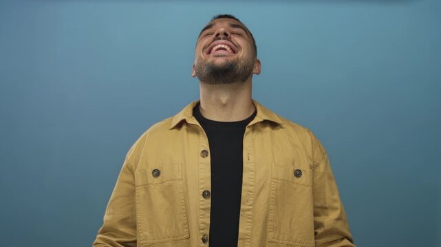 Young hispanic man in mustard jacket covers eyes with hands against blue studio wall in moody pose; playfulness joy whimsy delight.