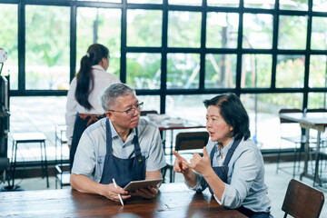 Elderly asian couple in aprons sitting at wooden table discussing cafe sales report using tablet...