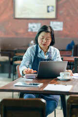 Asian middle aged female cafe owner wearing apron working on laptop with documents and coffee cup...