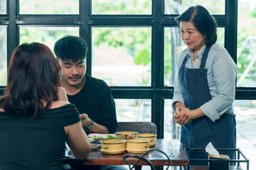 Asian senior female restaurant owner in apron standing near couple table observing and ensuring good service hospitality teamwork and customer satisfaction reflecting small business management concept