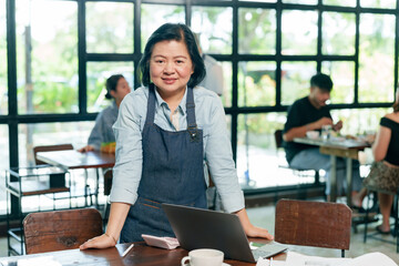 Asian elderly female restaurant owner standing near laptop smiling confidently in cafe representing...