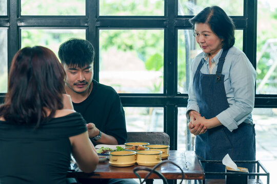 Asian senior female restaurant owner in apron standing near couple table observing and ensuring good service hospitality teamwork and customer satisfaction reflecting small business management concept