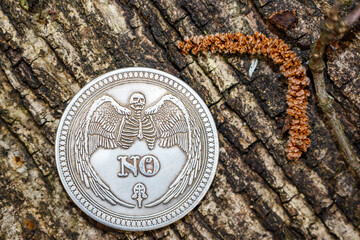 Gothic silver coin showcasing a winged skeleton and 'NO' inscription resting on rugged tree bark. A...