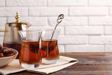 Tasty Turkish tea in glass cups, dates and teapot on wooden table against white brick wall, closeup. Space for text