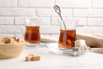 Tasty Turkish tea in glass cups, brown sugar and sweets on white marble table against brick wall, closeup