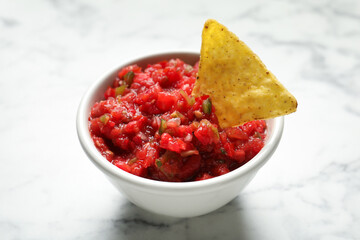 Tasty salsa in bowl and nacho on white marble table, closeup
