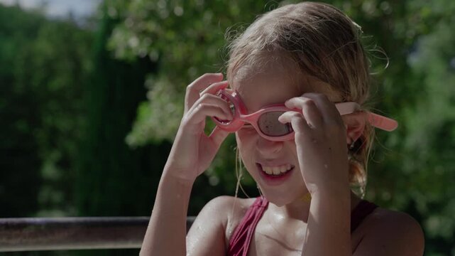 Young girl in a red swimsuit adjusting pink swimming goggles on a sunny day with green trees in the background