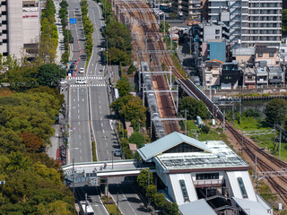 Aerial view of an Osaka district shows rail tracks, an elevated station with a pale green roof, a tree lined boulevard, and parks near Osaka Castle Park in crisp light.
