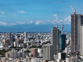 Obraz premium Central Tokyo, Japan, spreads with mid and high rises, cranes mark towers under construction, a cylindrical glass tower stands, blue mountains appear in a clear afternoon.